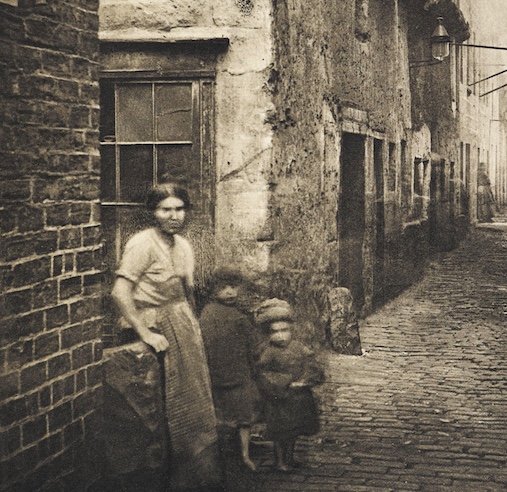 A poor woman with two bare foot children in a very run down street. The street is paved with cobbles.